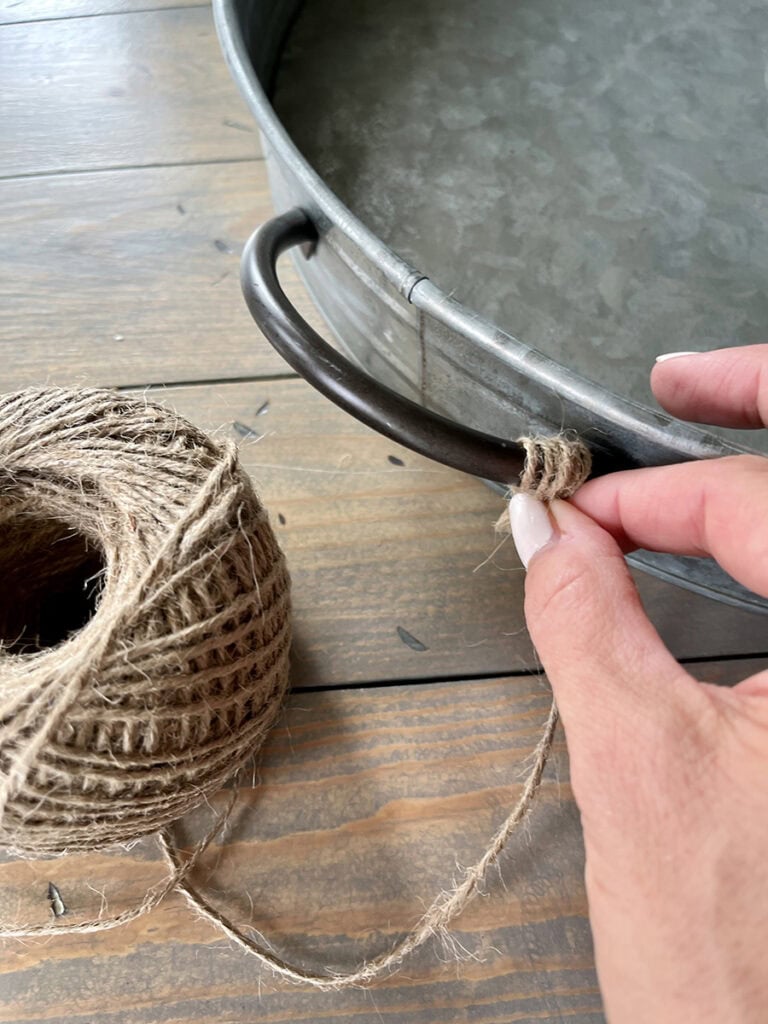 A close-up of a hand wrapping twine around the handle of a galvanized tray, with a spool of twine in the foreground.