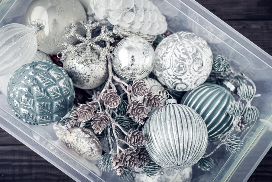 A plastic storage bin filled with silver, blue, and white Christmas ornaments and pinecones neatly organized by color.