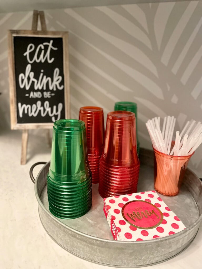 A galvanized tray holding red and green plastic cups, polka-dot napkins with “Be Merry” text, and a cup of white straws, with a festive “Eat, Drink, and Be Merry” sign in the background.