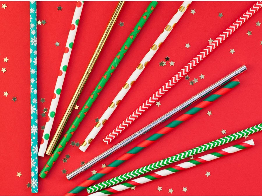 A colorful assortment of festive paper straws with holiday patterns, arranged on a red background sprinkled with gold star confetti to build a festive cocktail station.