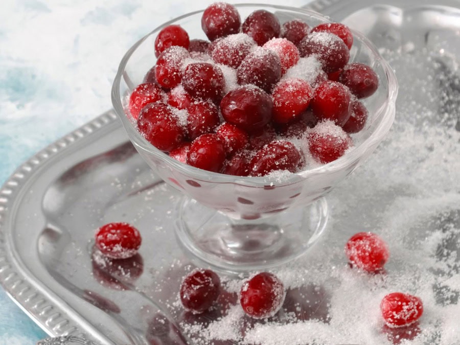 A clear glass dish filled with sugared cranberries, resting on a silver tray with a dusting of sugar around it.