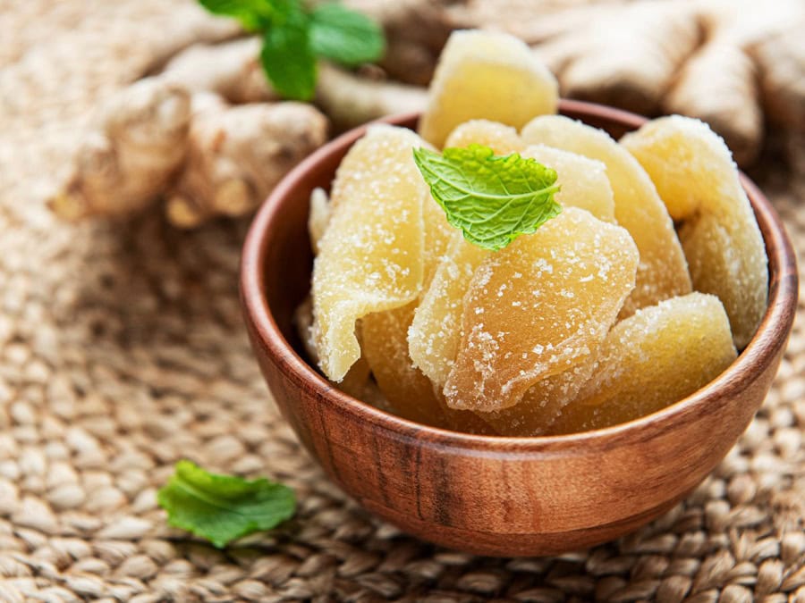 A wooden bowl filled with sugared candied ginger slices, garnished with a fresh mint leaf, set on a natural woven mat.