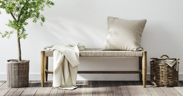 A woven bench styled in a bright, coastal entryway with a textured pillow, a cozy blanket, a wicker basket, and a potted plant nearby.