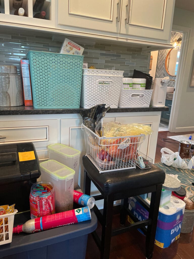 A cluttered kitchen counter and stool covered with baskets, pantry items, and storage bins during a pantry organization project.