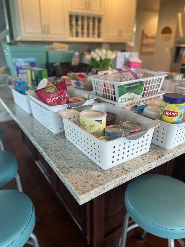 A kitchen island covered with white storage baskets filled with canned goods, dry pantry staples, and other food items during a pantry reorganization.
