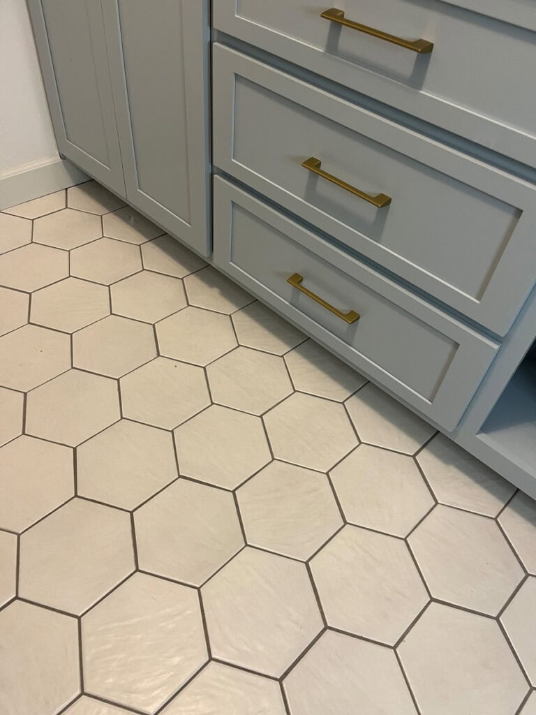 Light-colored hex tile flooring with dark grout paired with soft blue cabinets and gold hardware in a laundry room makeover, creating a modern yet timeless look.