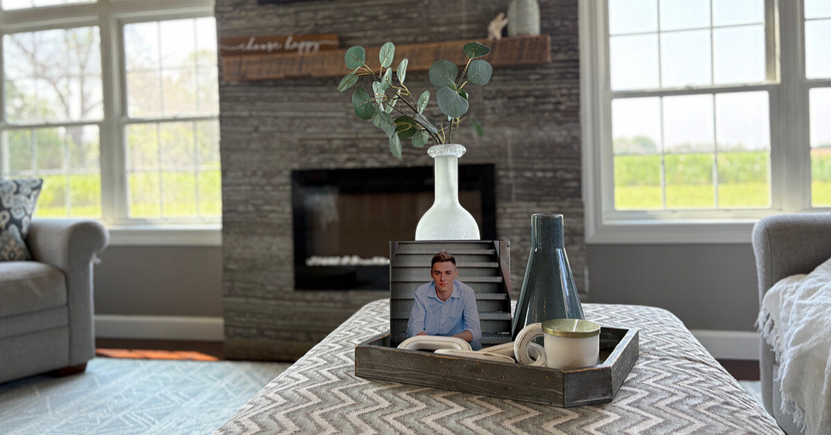 Coastal coffee table book decor with propped photo book, eucalyptus stem, vases, and candle styled in a wood tray on a patterned ottoman.