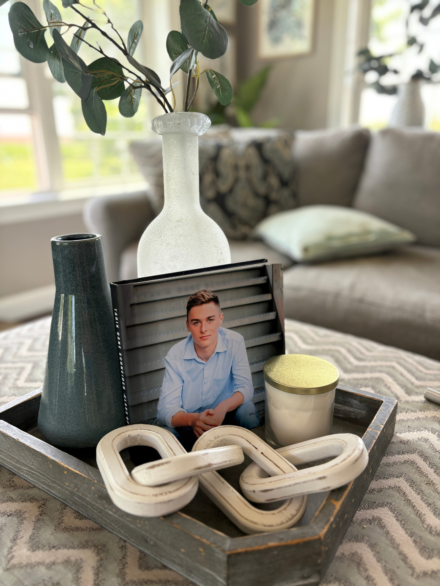 Styled tray with coffee table book decor featuring a senior photo album, eucalyptus stem, candle, and wood chain accent in a coastal-inspired living room.
