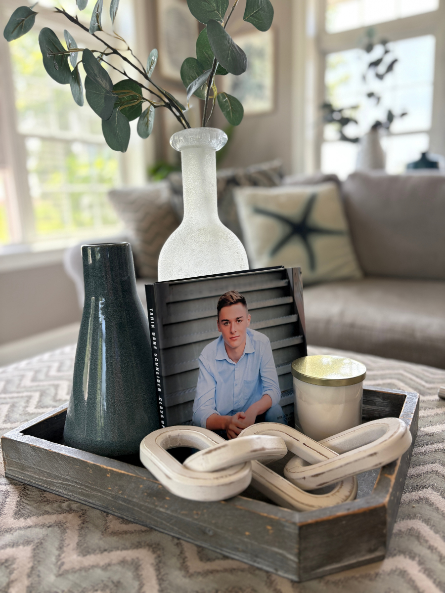 Coastal coffee table book decor with a senior photo book, eucalyptus stem, candle, and ceramic vases styled in a gray tray.