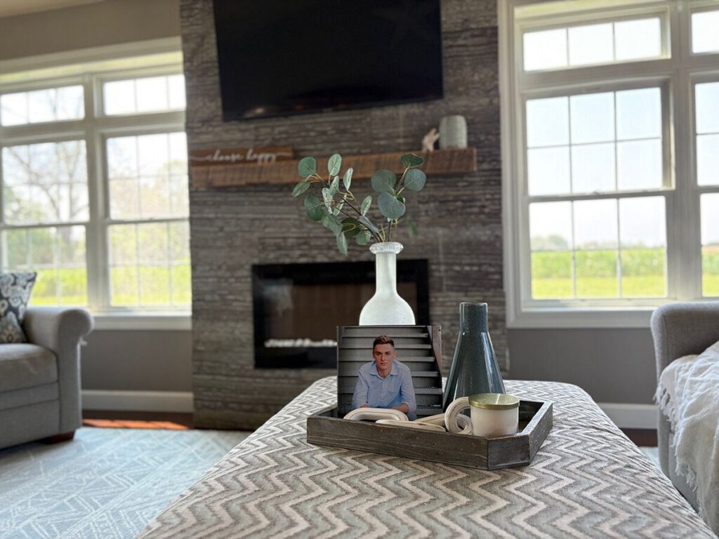 Coastal coffee table book decor setup on an upholstered ottoman with a wood tray, photo album, eucalyptus stem, ceramic vases, candle, and decorative accent.
