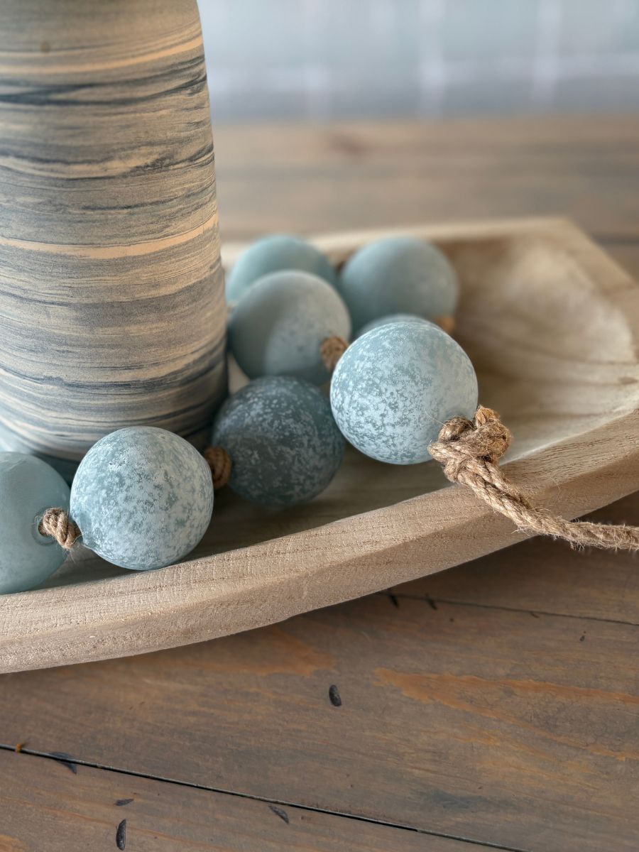 Close-up of a blue glass beaded garland looped around a marbled coastal-style vase in a carved wood tray.