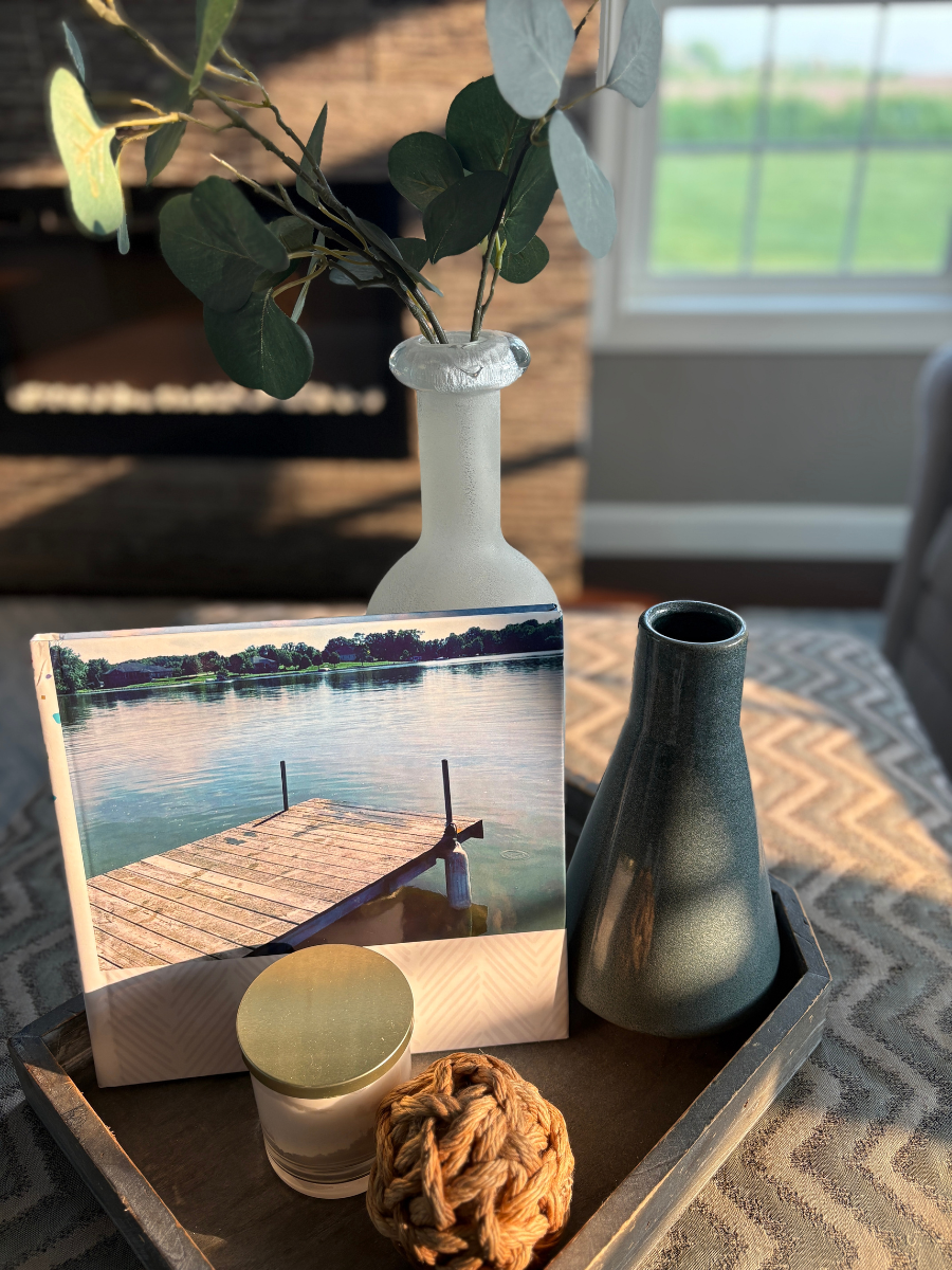 Coastal coffee table book decor featuring a photo book with a pier image, frosted vase with eucalyptus, green ceramic vase, candle, and nautical rope accent in a wood tray.
