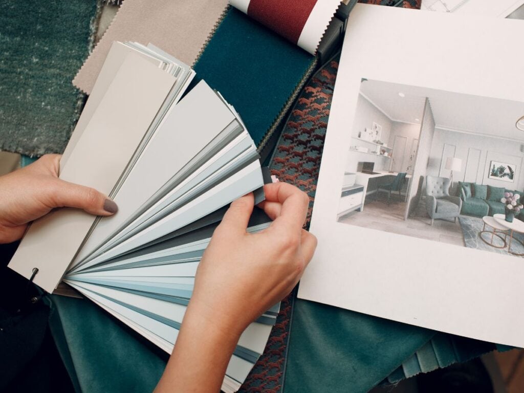 Hands holding a fan deck of interior paint color swatches alongside fabric samples and a styled mood board