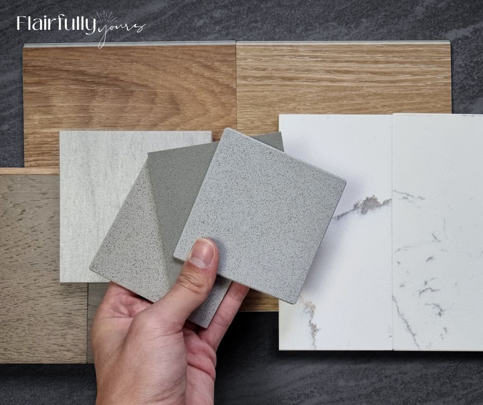 Hand holding gray countertop samples over wood flooring and quartz surfaces — comparing finishes for a home renovation checklist.