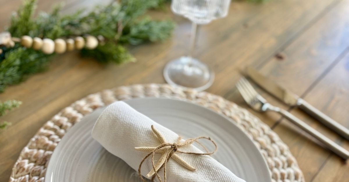 Simple Thanksgiving table decor with woven placemat, rolled linen napkin tied with jute and starfish, and greenery garland — a relaxed coastal tablescape idea for busy hosts.
