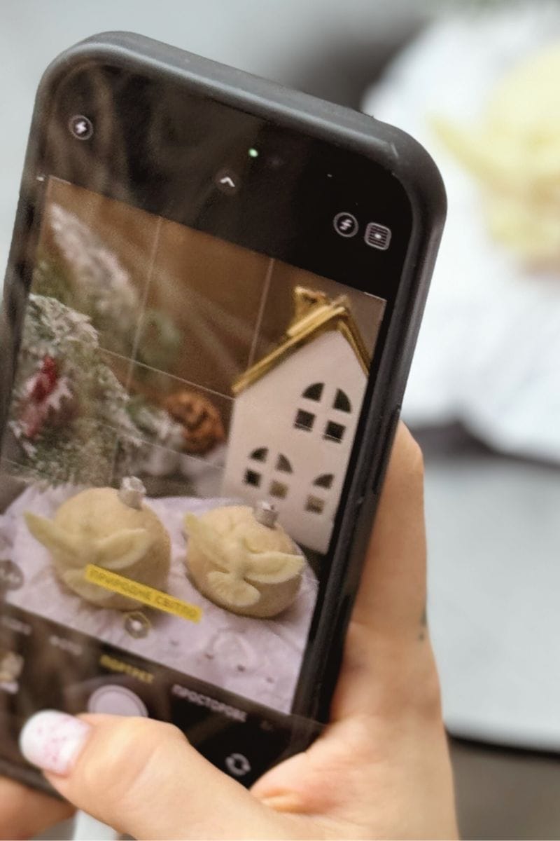 Close-up of a phone capturing a photo of tabletop Christmas décor, including two angel ornaments and a ceramic house with gold trim.