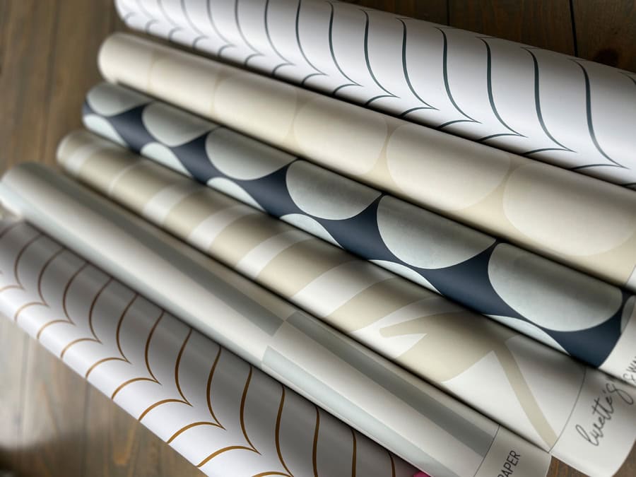 A stack of wallpaper rolls from Livette's, featuring various neutral and bold coastal prints, including a palm design, arranged neatly on a wooden surface.