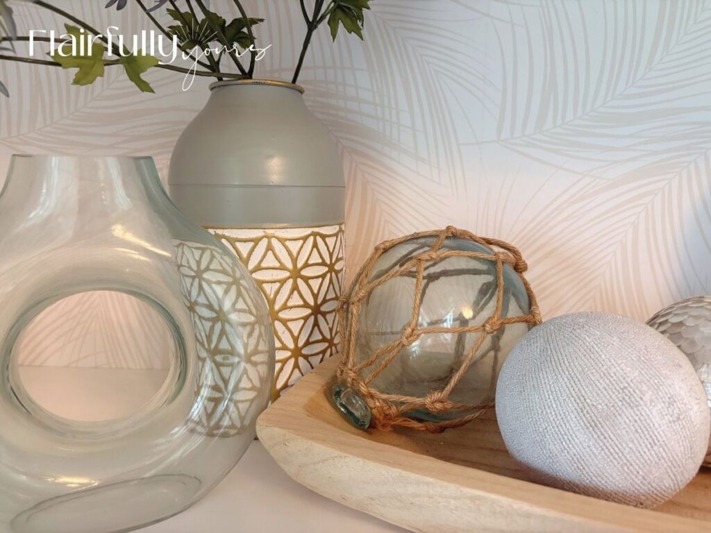 Close-up of a shelf featuring coastal decor with palm print wallpaper as the backdrop. Includes woven vases, a glass float ball, and a tall grass plant.