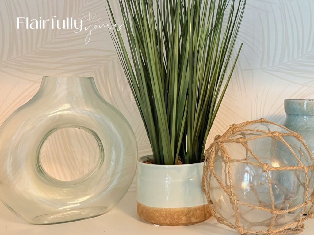 A wide shot of a shelf decorated with coastal accents, including glass vases, a framed picture, and a potted plant, all against a palm print wallpaper backdrop.