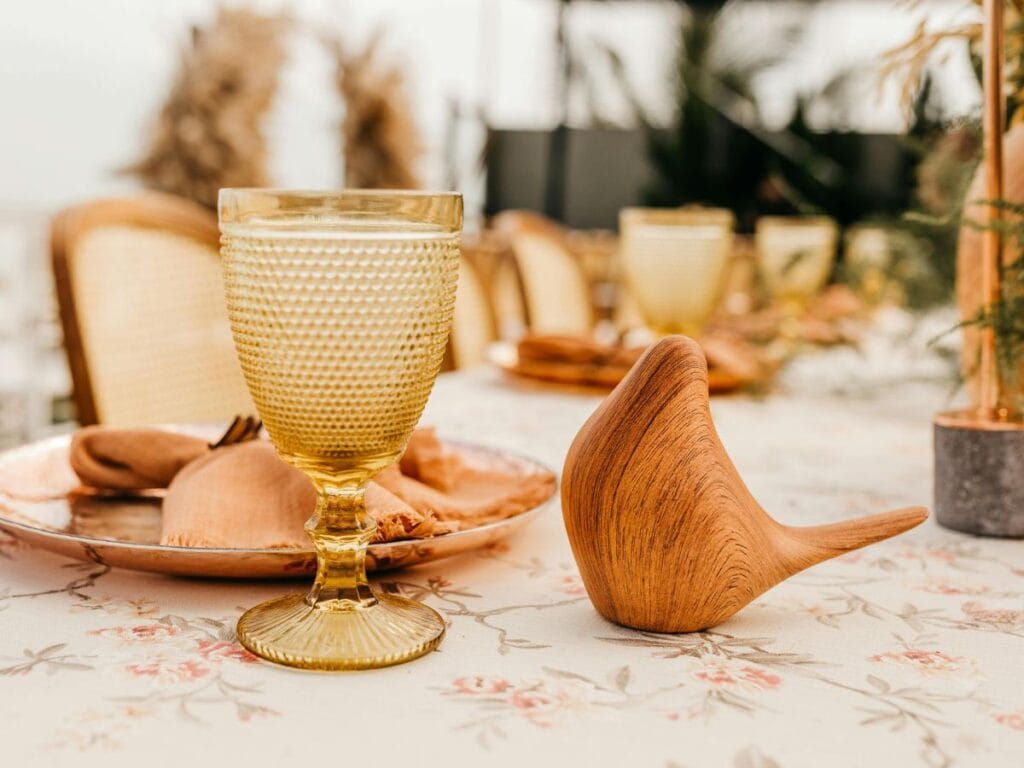 Table with amber glassware, wooden bird sculpture, and neutral tablecloth, illustrating tonal layering in a small-scale vignette.