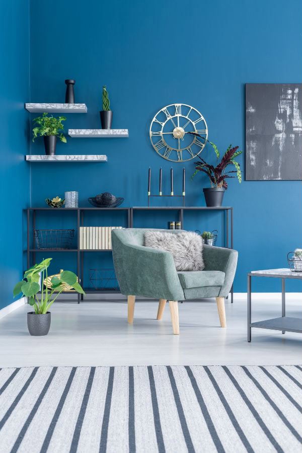 A living room with bold, room-wide tonal color: teal armchair, deep blue walls, and decorative accents in dark and medium blue hues, with gold and marble shelves holding plants and décor.