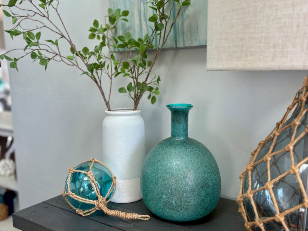 Three decorative items —aqua décor items on a table — a teal glass float, medium aqua vase, and deep turquoise ceramic vase — next to the edge of a lamp and corner of artwork, showing tonal variation and textures