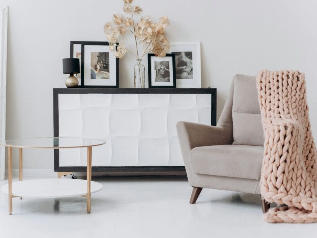 Modern living room corner with a neutral armchair draped with a chunky blush knit throw, a white wavy-front console topped with black-and-white framed photos, a gold-accented black lamp, and dried metallic eucalyptus in a clear vase, paired with a round glass-and-gold coffee table.