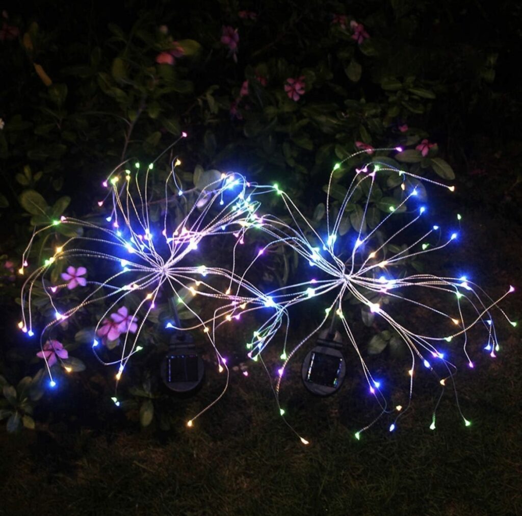 Solar “firework” garden lights placed in an outdoor planter with colorful summer flowers, glowing at night, adding height, texture, and sparkle to the arrangement.
