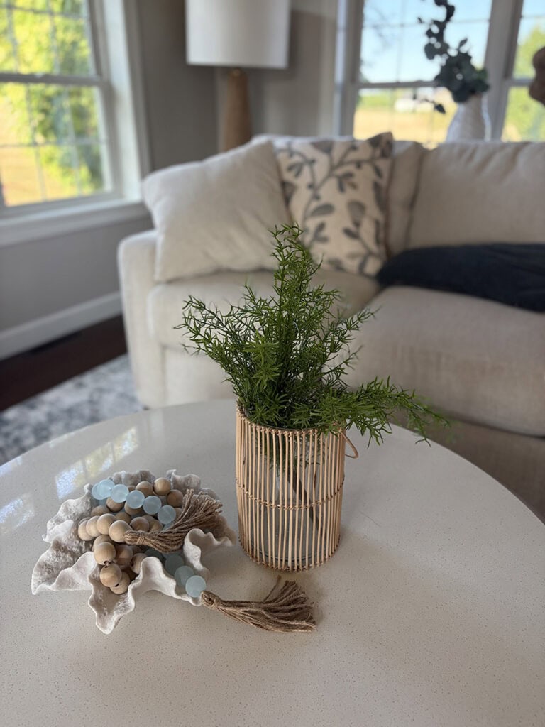 Coastal round coffee table decor featuring an aqua bead garland in a coral bowl beside a woven vase filled with greenery.