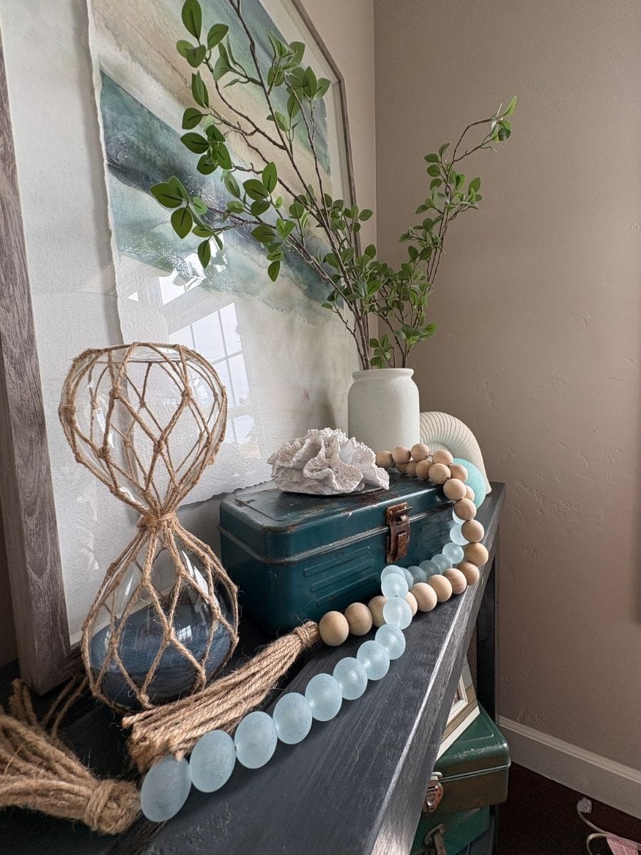 Rope-wrapped glass hourglass adding height to a coastal console table with greenery, sea glass beads and coral