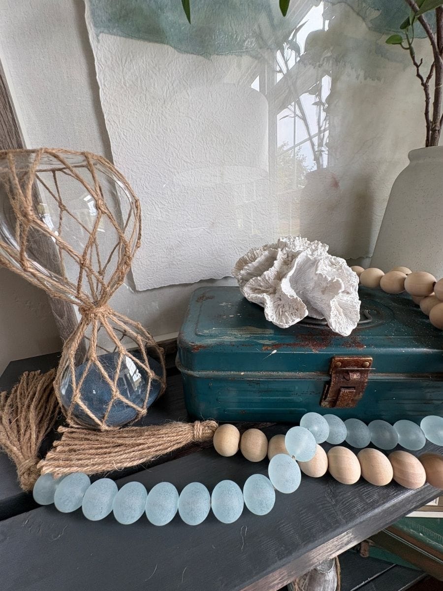 Wood and aqua sea glass bead garland draped across a coastal console table with white coral and rope-wrapped glass hourglass