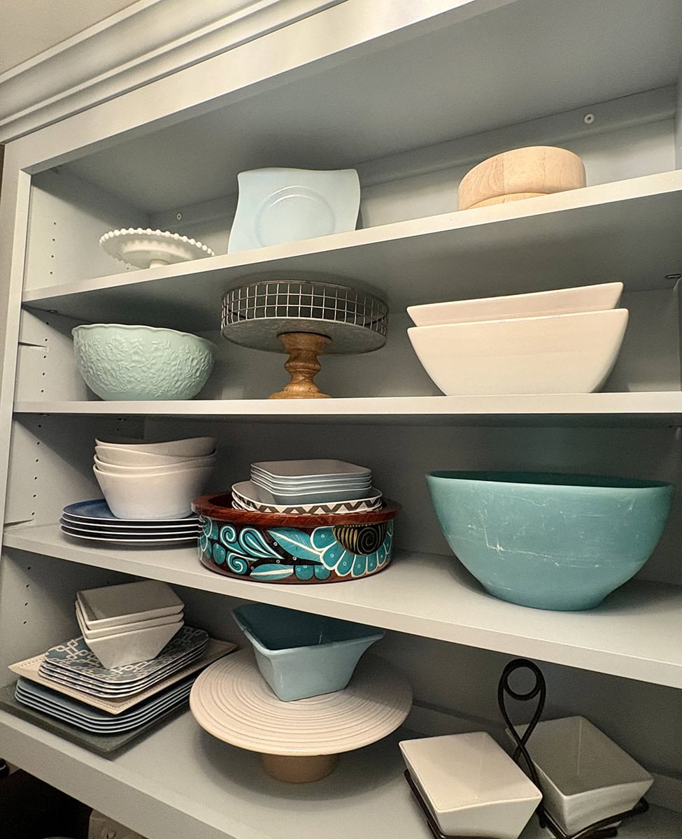 Pantry shelves filled with white serving dishes, aqua bowls, glass pieces and a colorful statement serving bowl.