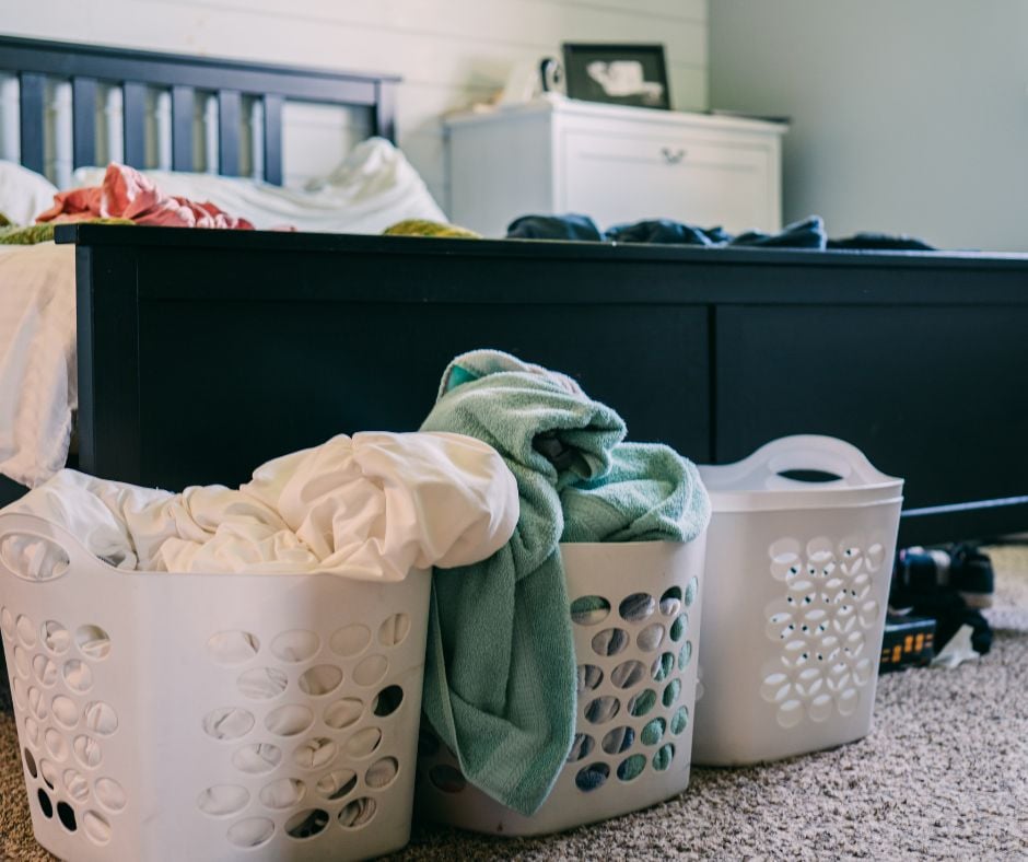 Three white laundry baskets filled with clothes and towels on a carpeted bedroom floor, with additional clothing scattered across an unmade bed.