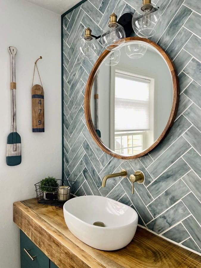 Round wood-framed bathroom mirror reflecting a bright window, surrounded by blue-gray herringbone tile, brass fixtures and a white vessel sink.