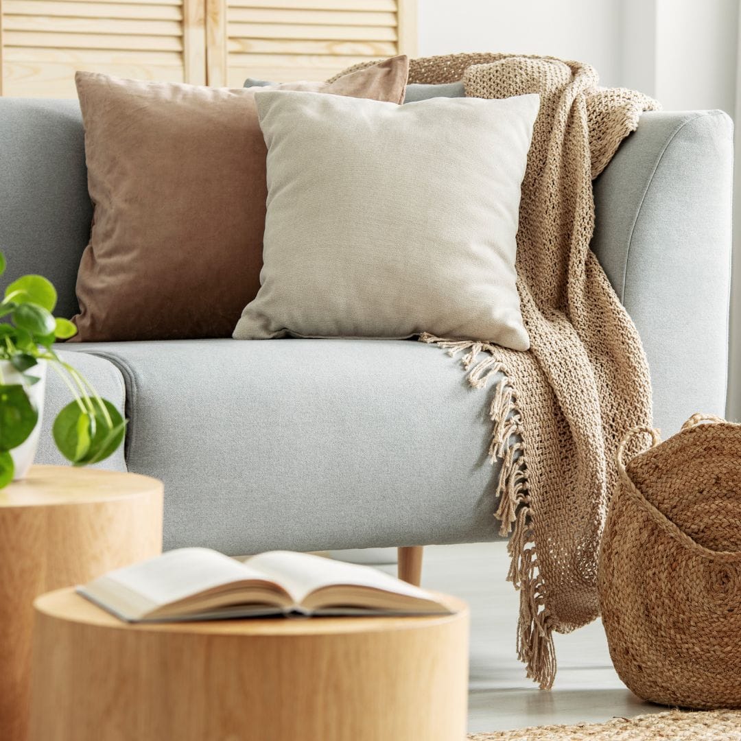 Chunky beige knit throw blanket with fringe draped over the arm of a light gray sofa with neutral decorative pillows and a woven basket.