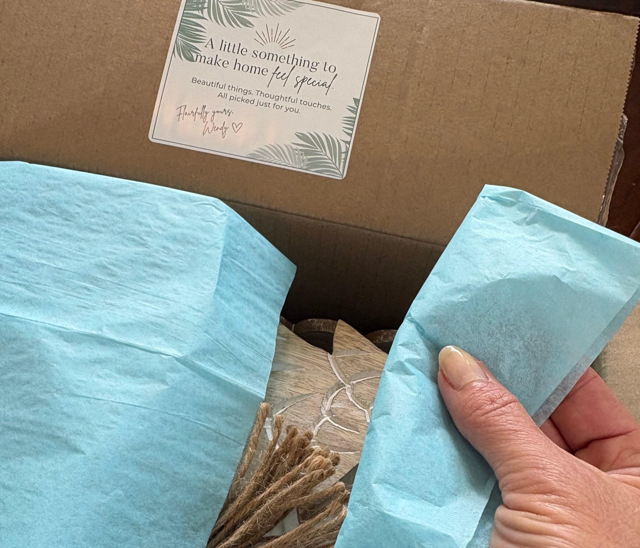 Hand lifting aqua tissue paper inside the Coastal Home Experience subscription box, revealing a glimpse of greenery, a white serving bowl, and wrapped home décor.