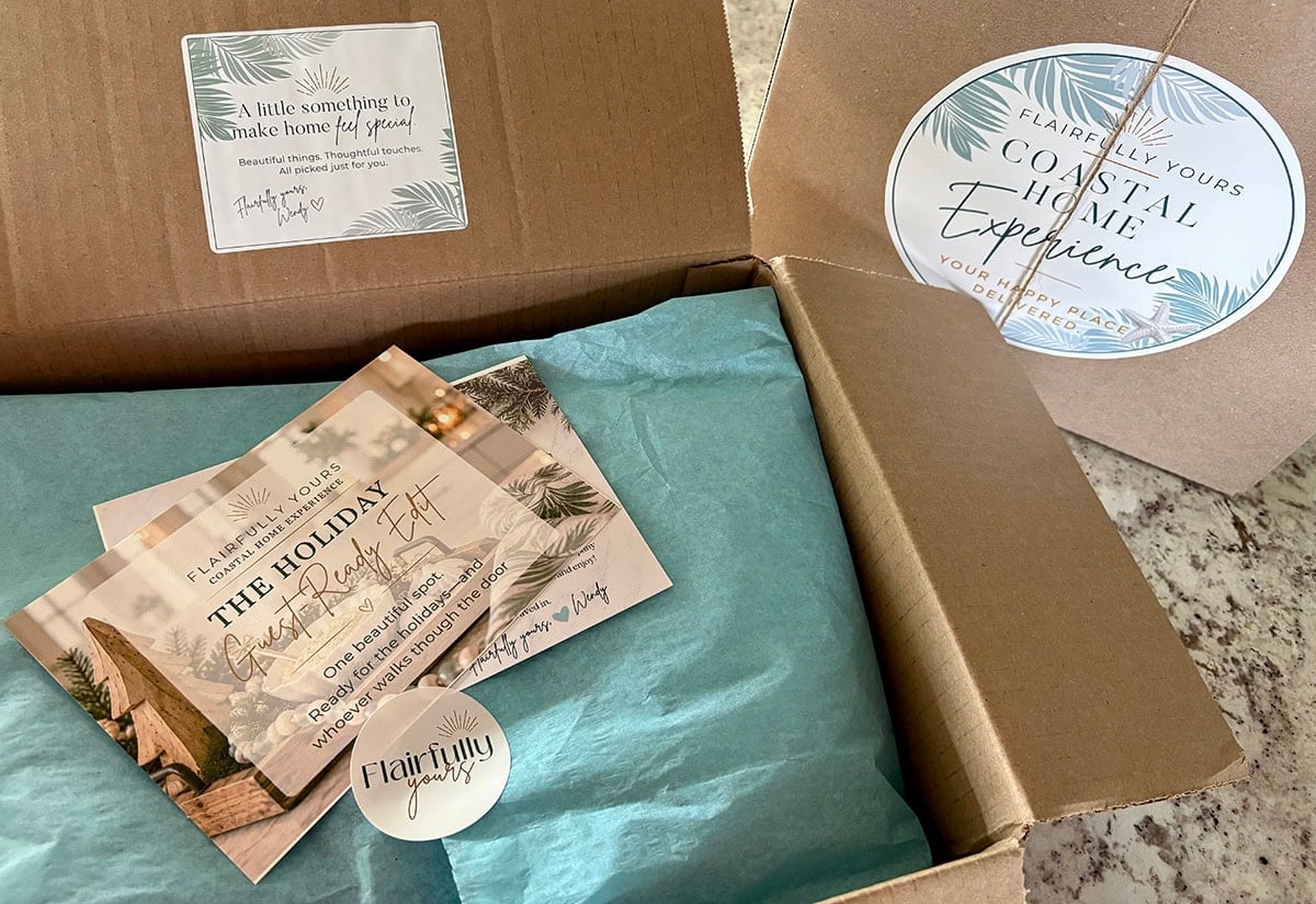 Open Flairfully Yours Coastal Home Experience subscription box with aqua tissue paper, branded packaging, and styling cards for the Holiday Guest-Ready Edit.